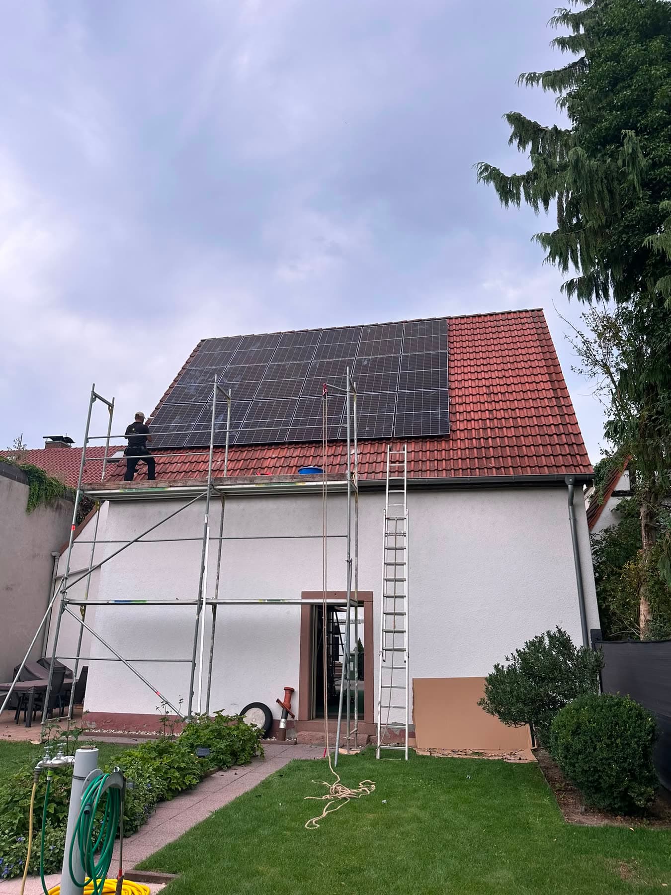 Eingerüstetes Wohnhaus, ein Monteur verlegt Solarmodule auf dem Ziegeldach